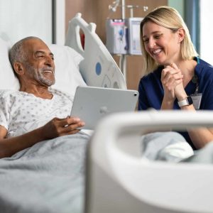 An older man in a hospital bed smiles while holding a tablet, with a nurse beside him, both enjoying the interaction—highlighting the positive impact of Apple Health Expansion in creating brighter, more connected hospital experiences.