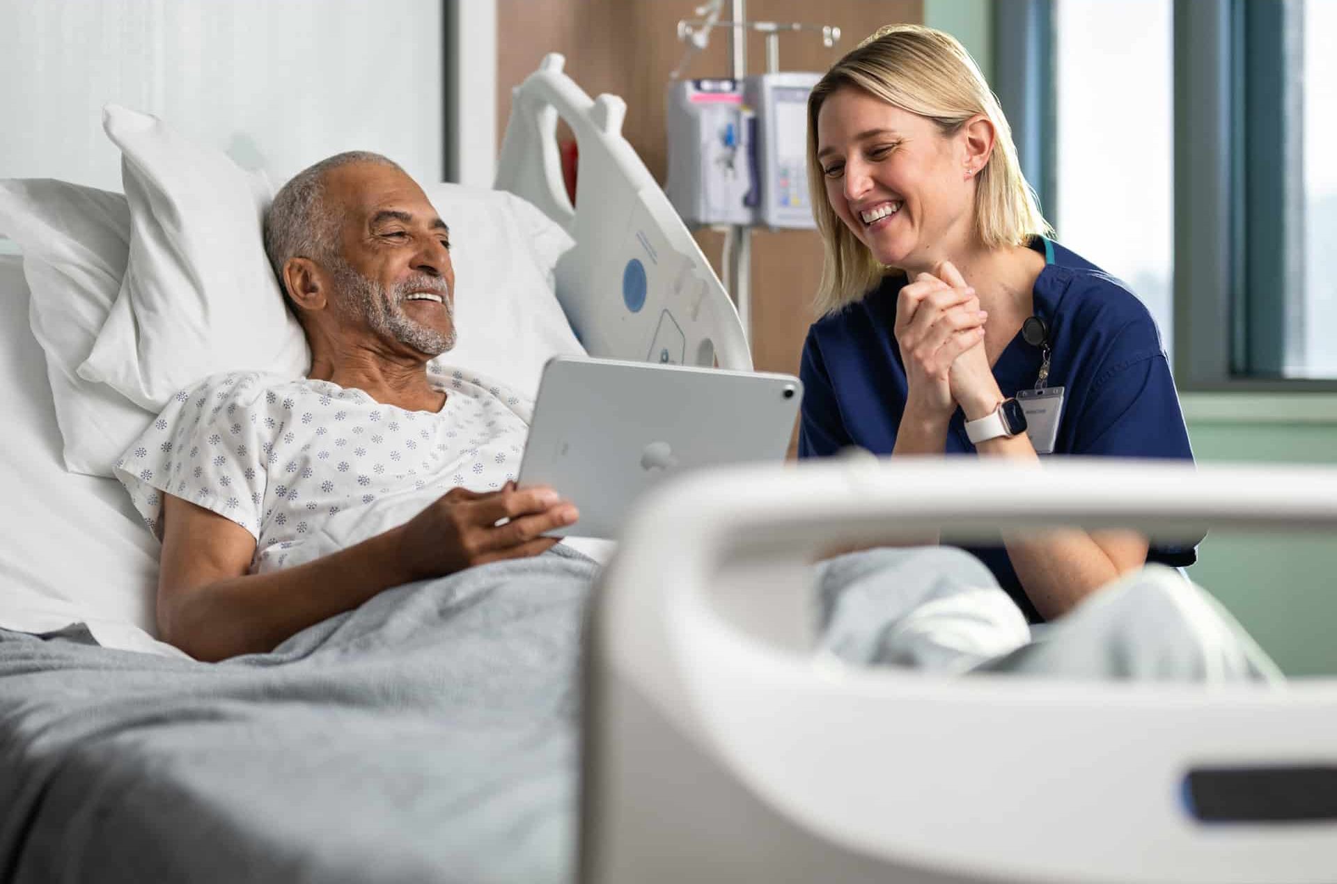 An older man in a hospital bed smiles while holding a tablet, with a nurse beside him, both enjoying the interaction—highlighting the positive impact of Apple Health Expansion in creating brighter, more connected hospital experiences.