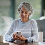 An older adult with gray hair, wearing a light blue striped sweater, sits at a table using a smartphone with Apple accessibility features. A cup sits nearby. She appears focused and content in a well-lit, cozy indoor setting.