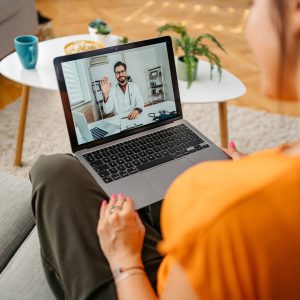 A woman sits on a sofa, holding a laptop on her lap in a remote therapy setup during a video call with a doctor. The doctor, wearing a white coat, is waving from the screen. A coffee table with mugs and snacks is in the background.