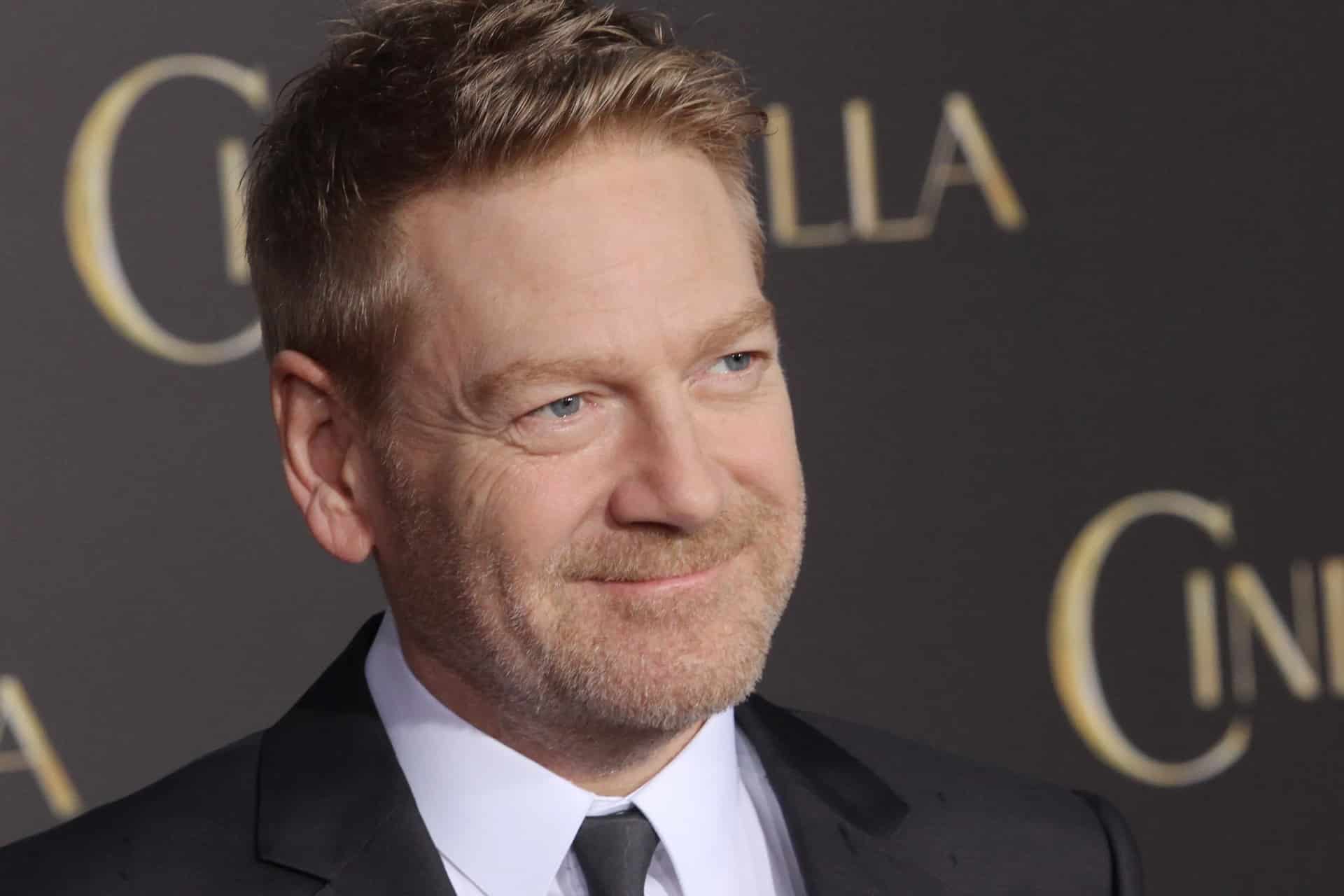 A man with light brown hair and a trimmed beard, dressed in a black suit and tie, smiles at an Apple TV Mayday event. The blurred background shows large gold letters on a dark backdrop.