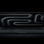 A sleek black MacBook Pro OLED is open on a dark surface, its screen displaying an abstract, glowing black and gray pattern of curved lines against a black background.