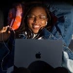 A young person with braided hair and glasses lies on a blanket outdoors, smiling with eyes closed, wearing earphones and holding a phone while a laptop rests on their chest—capturing the essence of teenage tech preference. Sunlight illuminates their face.