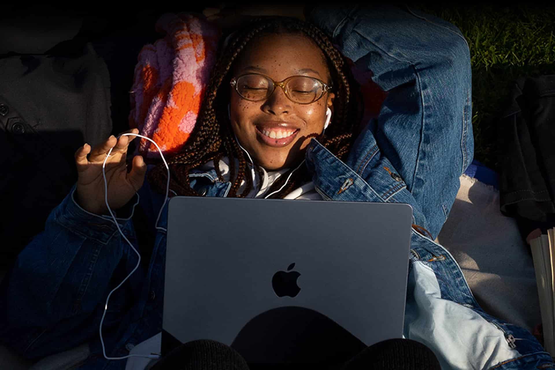 A young person with braided hair and glasses lies on a blanket outdoors, smiling with eyes closed, wearing earphones and holding a phone while a laptop rests on their chest—capturing the essence of teenage tech preference. Sunlight illuminates their face.