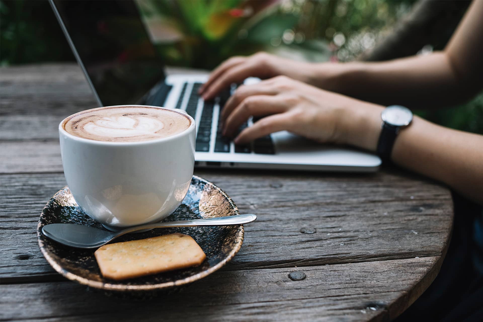 A close-up of a cappuccino with latte art and a biscuit on a saucer, set on a wooden table. In the background, a person works in their Remote Office, typing on a laptop with greenery behind them.