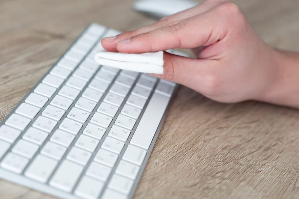 A person cleans a white Magic Keyboard with a folded white cloth on a wooden desk.