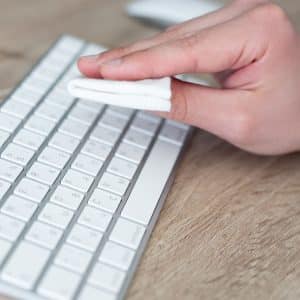 A person cleans a white Magic Keyboard with a folded white cloth on a wooden desk.