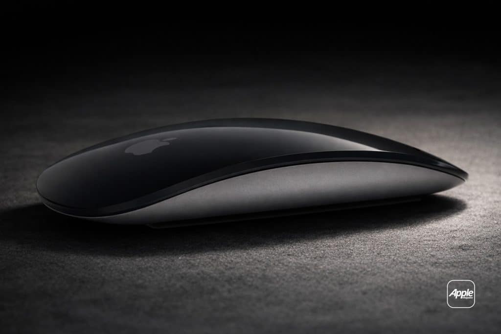 A sleek, black Apple Magic Mouse rests on a dark surface, illuminated by soft lighting. The Apple logo is visible on the mouse, highlighting its intuitive Magic Mouse Gestures. An Apple-branded badge appears in the lower right corner.