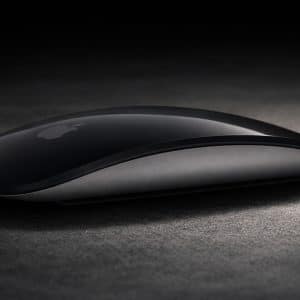 A sleek, black Apple Magic Mouse rests on a dark surface, illuminated by soft lighting. The Apple logo is visible on the mouse, highlighting its intuitive Magic Mouse Gestures. An Apple-branded badge appears in the lower right corner.