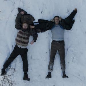 Two men lie on their backs in the snow, arms outstretched, looking up. One wears a vest and sweater, the other a jacket and shirt. Footprints and tree branches are visible around them in this Apple TV Mayday scene.