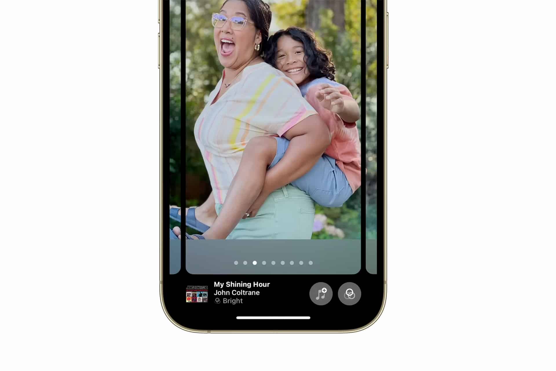 A smartphone screen shows a photo of a smiling woman giving a piggyback ride to a laughing child outdoors. At the bottom, music by John Coltrane titled “My Shining Hour” plays, capturing the joy of Photo Memory Videos.