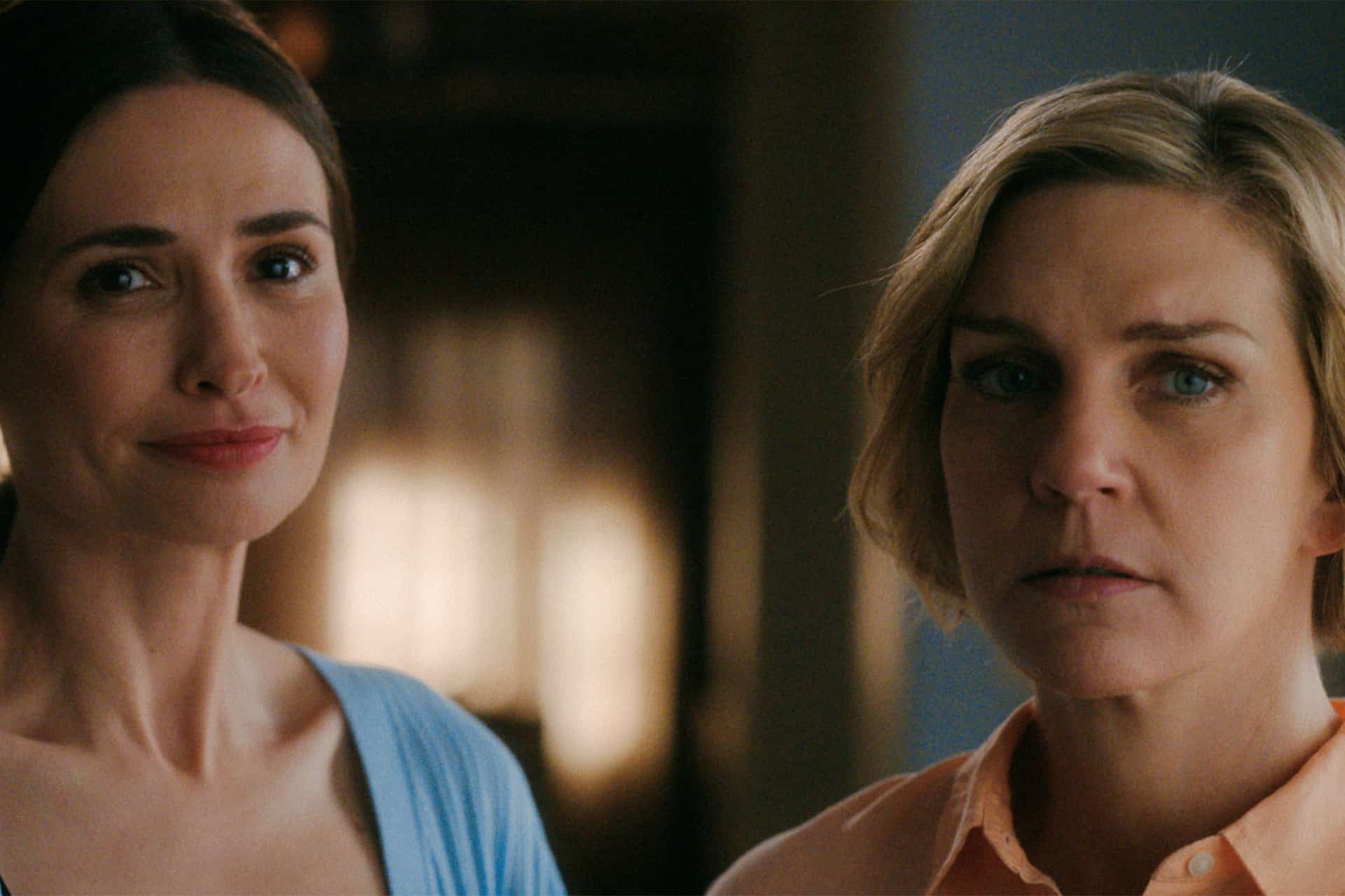 Two women stand side by side indoors in a scene from Pluribus Season 2; one has long dark hair with a faint smile, while the other sports short blonde hair and a serious expression. Warm lighting creates a soft background.