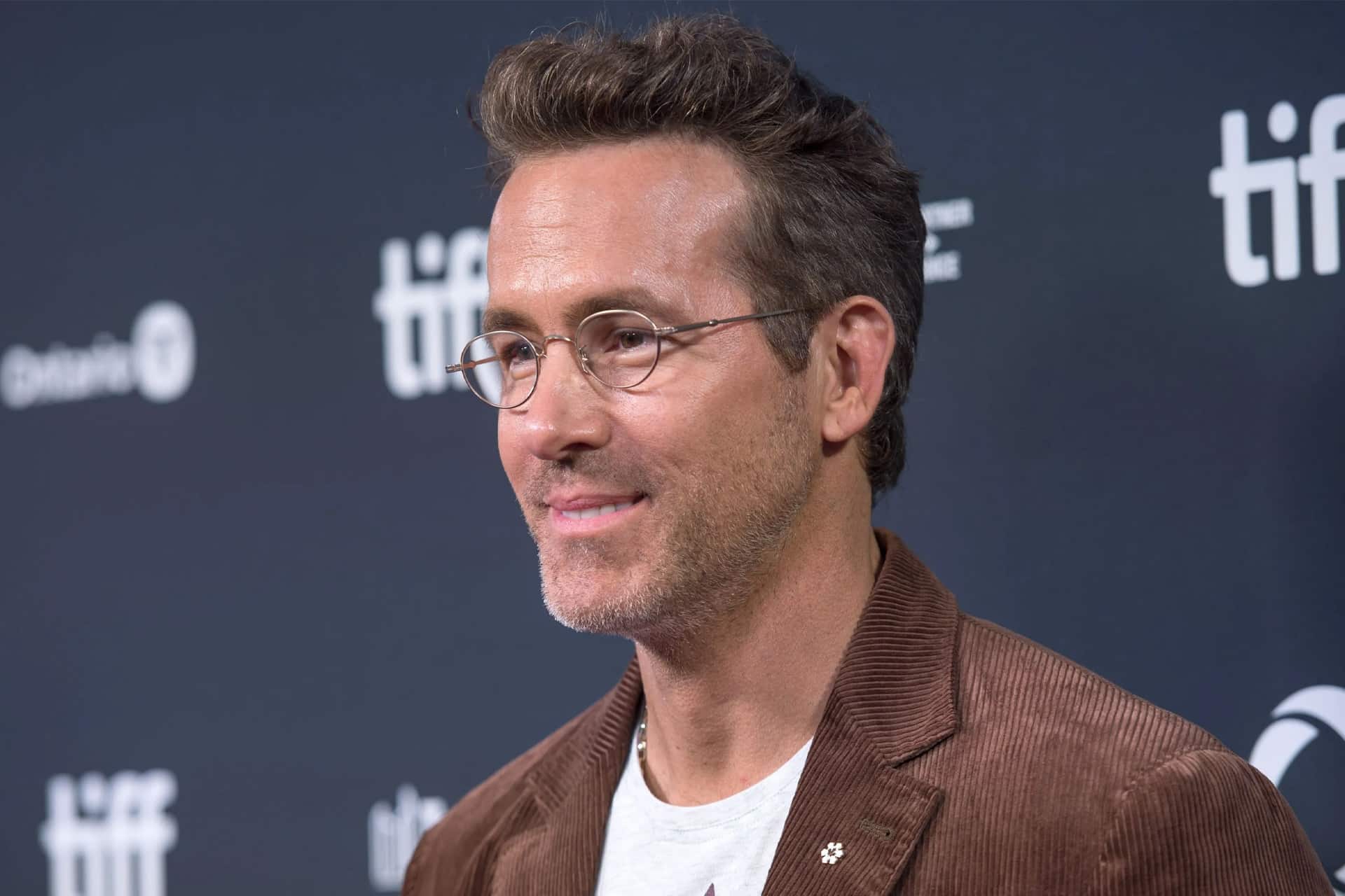 A man with short brown hair and glasses, wearing a brown jacket and white shirt, smiles in front of a dark backdrop with "tiff" logos, possibly attending an Apple TV Mayday event.