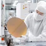 A person in a cleanroom suit and gloves examines a silicon wafer in a high-tech lab, showcasing the precision and innovation of Apple US Production, with advanced equipment and machinery visible in the background.