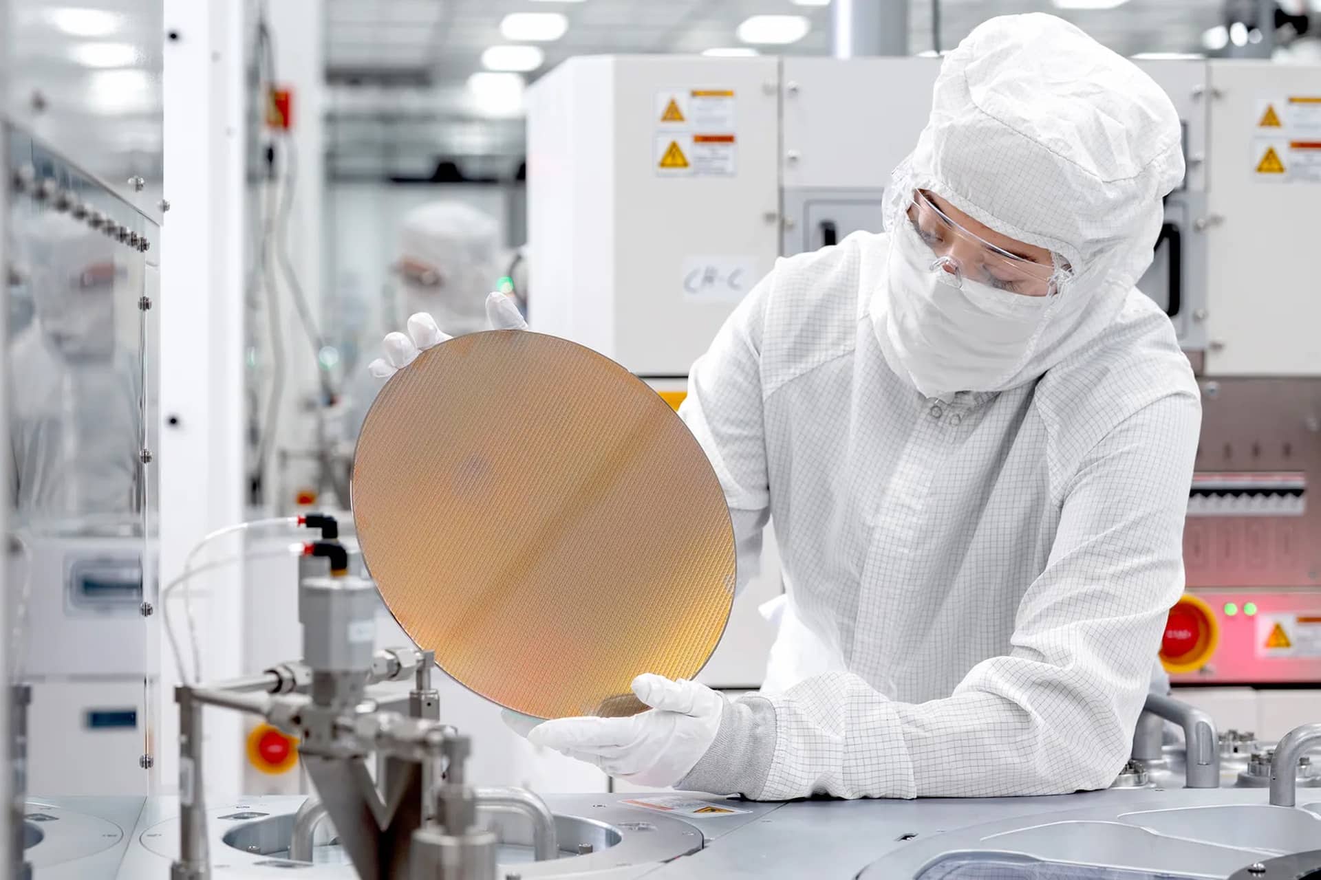 A person in a cleanroom suit and gloves examines a silicon wafer in a high-tech lab, showcasing the precision and innovation of Apple US Production, with advanced equipment and machinery visible in the background.
