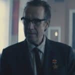 A middle-aged man in glasses and a suit stands in a dimly lit room, wearing two medals on his lapel—a scene reminiscent of the political intrigue found in Apple TV’s For All Mankind. Blurred seats and red flags hang in the background.