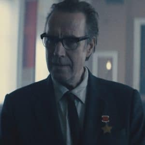 A middle-aged man in glasses and a suit stands in a dimly lit room, wearing two medals on his lapel—a scene reminiscent of the political intrigue found in Apple TV’s For All Mankind. Blurred seats and red flags hang in the background.