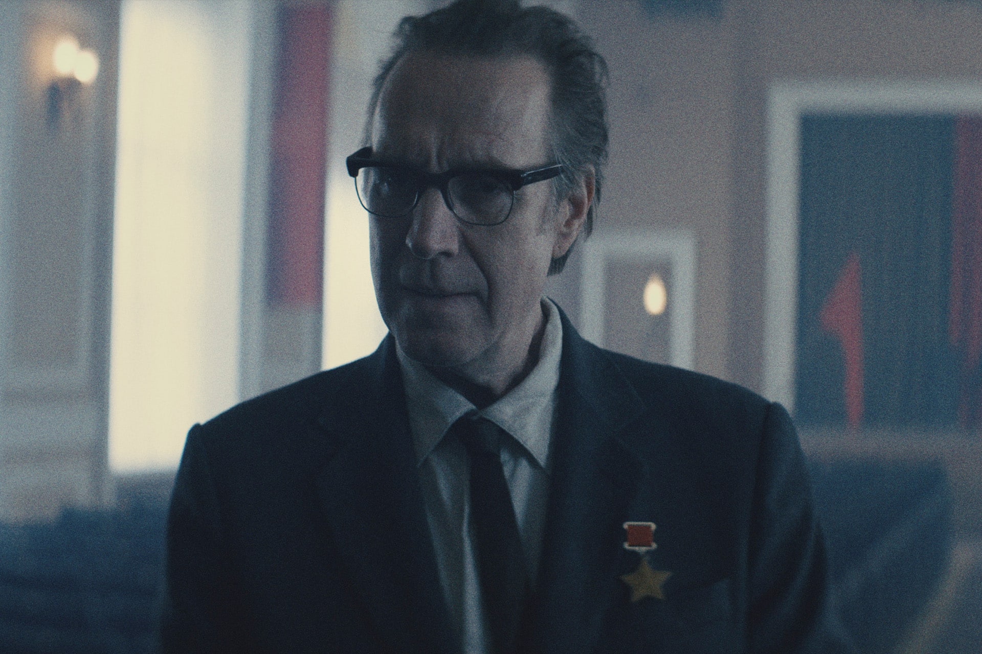 A middle-aged man in glasses and a suit stands in a dimly lit room, wearing two medals on his lapel—a scene reminiscent of the political intrigue found in Apple TV’s For All Mankind. Blurred seats and red flags hang in the background.