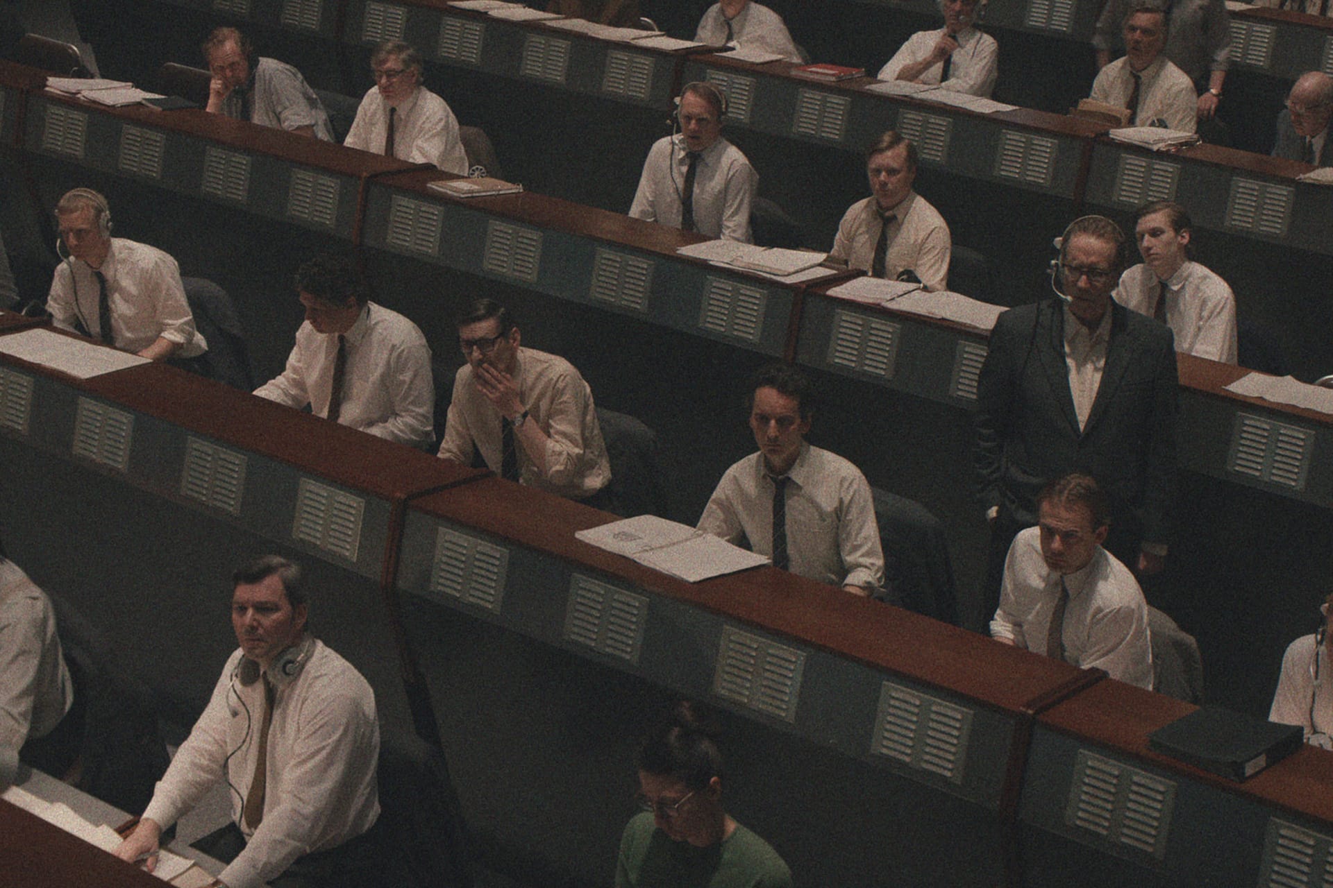 A room filled with men in white shirts and ties sitting at desks with headsets and papers, resembling a 1960s mission control center—like a scene straight out of For All Mankind on Apple TV.