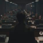 A dimly lit office with rows of women at typewriters exudes an orderly, serious atmosphere—evoking scenes reminiscent of For All Mankind on Apple TV. A solitary figure stands facing them in the foreground, viewed from behind.