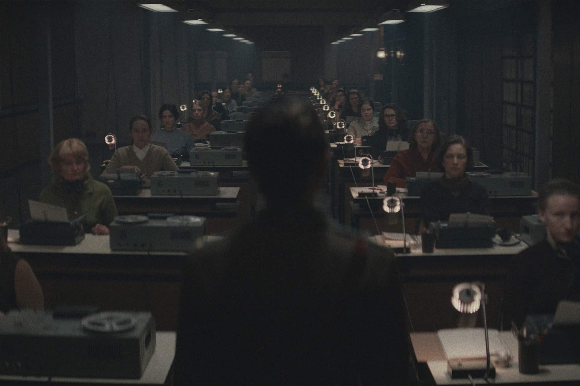 A dimly lit office with rows of women at typewriters exudes an orderly, serious atmosphere—evoking scenes reminiscent of For All Mankind on Apple TV. A solitary figure stands facing them in the foreground, viewed from behind.