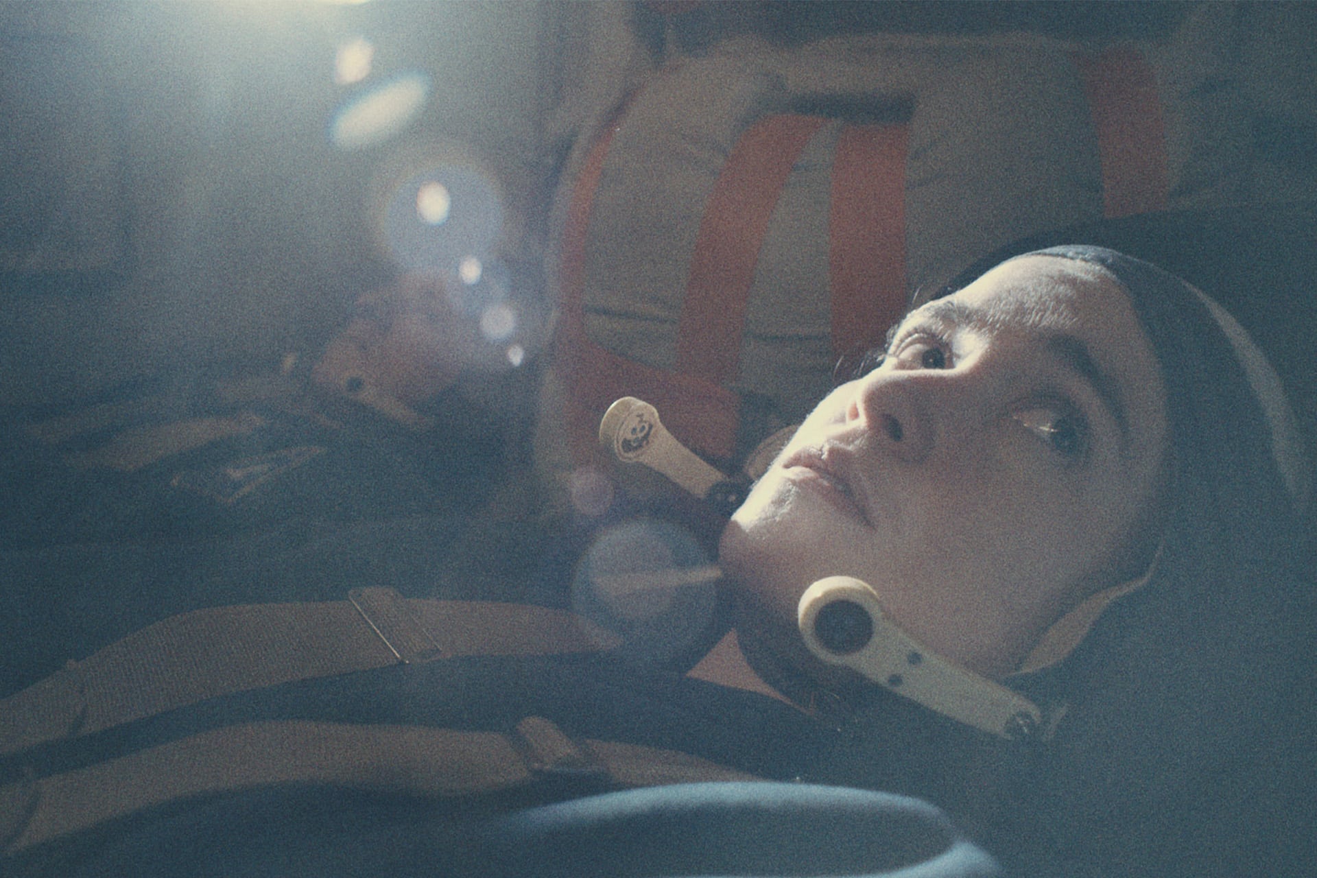 A person wearing a helmet and gear lies down, looking upward, with a soft light and lens flare illuminating their face. Another figure, similarly dressed, is blurry in the background. The muted, cinematic scene echoes Apple TV's For All Mankind.