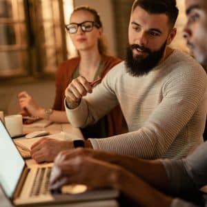 Three people are sitting at a desk in a Startup Garage, working on laptops. One man points at the laptop screen while talking to another man, as a woman with glasses observes. They appear to be collaborating on a project.