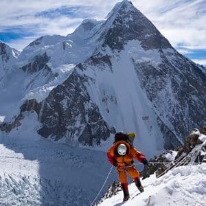 A climber in orange gear ascends a snowy, steep mountain slope using ropes, with majestic, snow-covered peaks and a glacier in the background—an adventure worthy of inspiring Apple documentaries—under a partly cloudy sky.