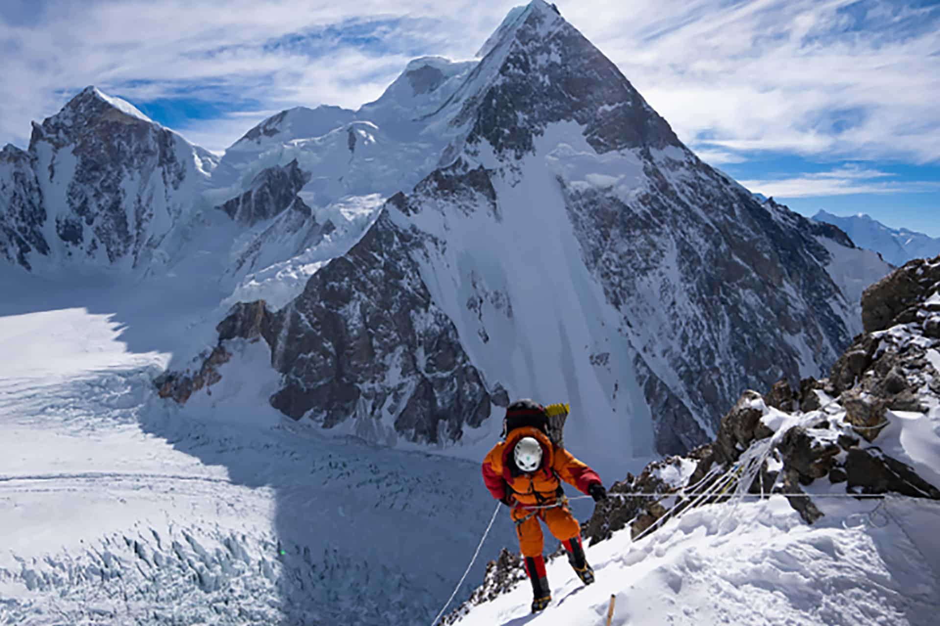 A climber in orange gear ascends a snowy, steep mountain slope using ropes, with majestic, snow-covered peaks and a glacier in the background—an adventure worthy of inspiring Apple documentaries—under a partly cloudy sky.