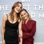 Two women smile and pose together in front of a wall with large text. One wears a black dress, and the other, in a burgundy suit, leans her head on her friend’s shoulder—like stars from an Apple TV series to watch.
