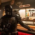 A person in black Mandalorian armor stands at a bar with Grogu on their shoulder, set in a warm, sci-fi Star Wars ambiance. Colorful bottles line the background, evoking the iconic Disney+ series.