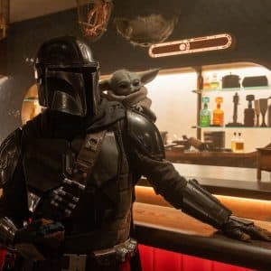 A person in black Mandalorian armor stands at a bar with Grogu on their shoulder, set in a warm, sci-fi Star Wars ambiance. Colorful bottles line the background, evoking the iconic Disney+ series.
