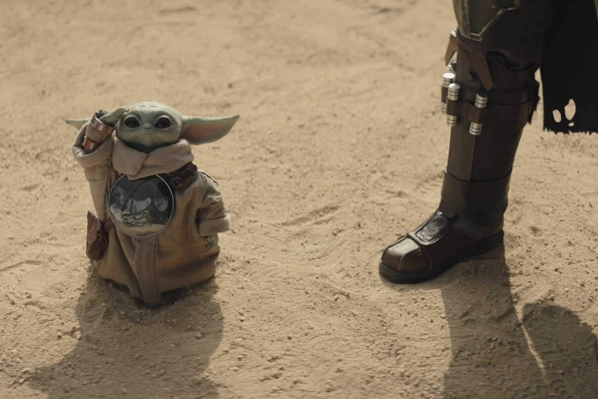 The Mandalorian and Grogu - A small alien creature with large ears and green skin stands in the sand next to a person wearing brown boots and armor. The Disney+ character is wearing a tan robe and looking upward.