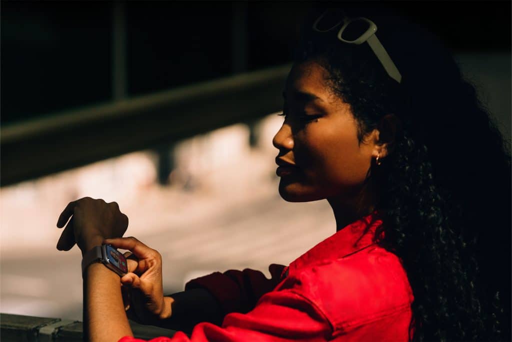 A woman in a red shirt stands in partial shadow, checking Apple Watch Gestures on her smartwatch. She has curly hair, sunglasses on her head, and is positioned in profile against a softly lit background.