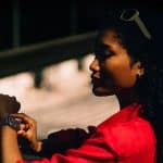 A woman in a red shirt stands in partial shadow, checking Apple Watch Gestures on her smartwatch. She has curly hair, sunglasses on her head, and is positioned in profile against a softly lit background.