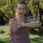 A woman with one arm, wearing a purple tank top and wireless earbuds, stands outdoors in a park looking at her smartwatch, about to use the Apple Watch Double Tap feature. Trees and greenery are blurred in the background.