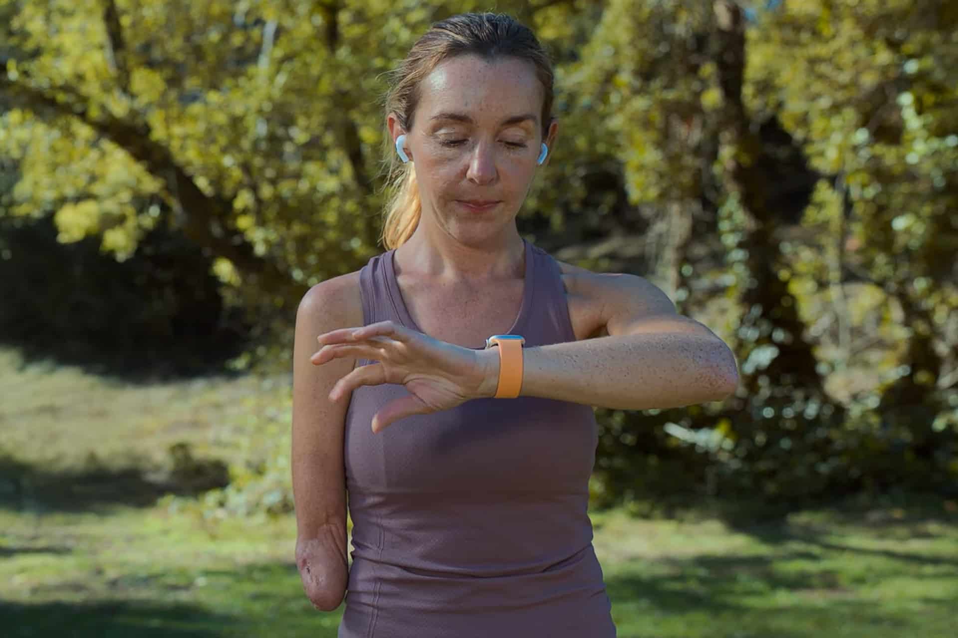 A woman with one arm, wearing a purple tank top and wireless earbuds, stands outdoors in a park looking at her smartwatch, about to use the Apple Watch Double Tap feature. Trees and greenery are blurred in the background.