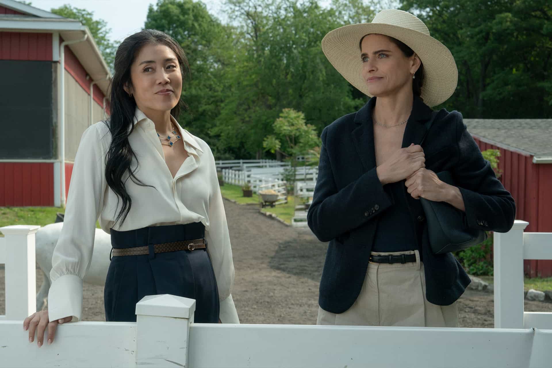 Two women stand by a white fence at a horse stable. One wears a white blouse and dark pants, while the other, ready for Apple TV’s "Your Friends & Neighbors" season 3, wears a hat, blazer, and holds a bag. Trees and a red barn are visible in the background.