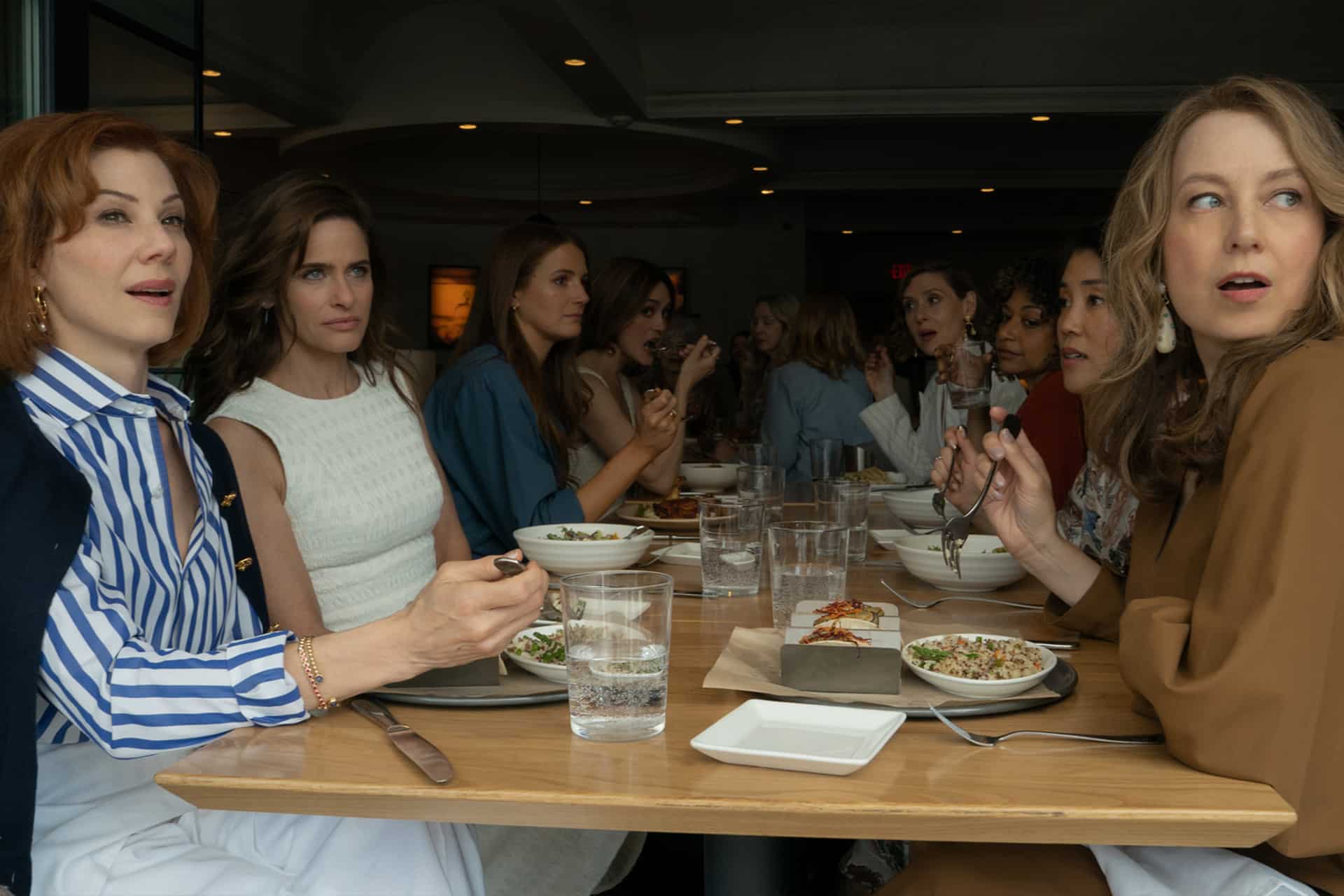 A group of women sit together at a restaurant table, eating and talking. Their serious, attentive expressions evoke the drama of Your Friends & Neighbors, now streaming season 3 on Apple TV. Plates of food, glasses, and utensils are on the table.