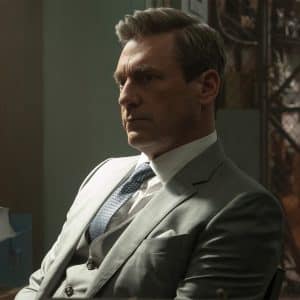 A man in a gray suit sits at a desk with stacks of papers, looking serious and deep in thought. The tense atmosphere and dim lighting evoke the drama of Your Friends & Neighbors, now streaming on Apple TV.