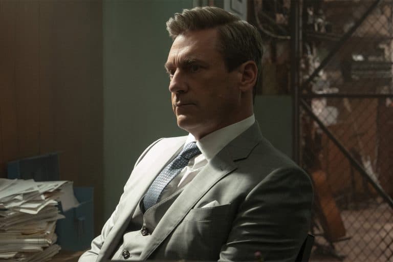 A man in a gray suit sits at a desk with stacks of papers, looking serious and deep in thought. The tense atmosphere and dim lighting evoke the drama of Your Friends & Neighbors, now streaming on Apple TV.