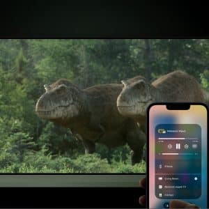 A person holds a smartphone using iPhone AirPlay to control media, while in the background a TV displays two realistic dinosaurs in a lush, green forest scene.