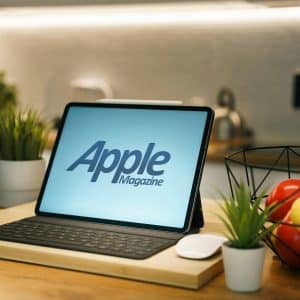 An iPad Kitchen companion with a keyboard and Apple Magazine on the screen sits on a wooden kitchen counter, surrounded by potted plants, a bowl of fruit, and kitchen appliances in the background.