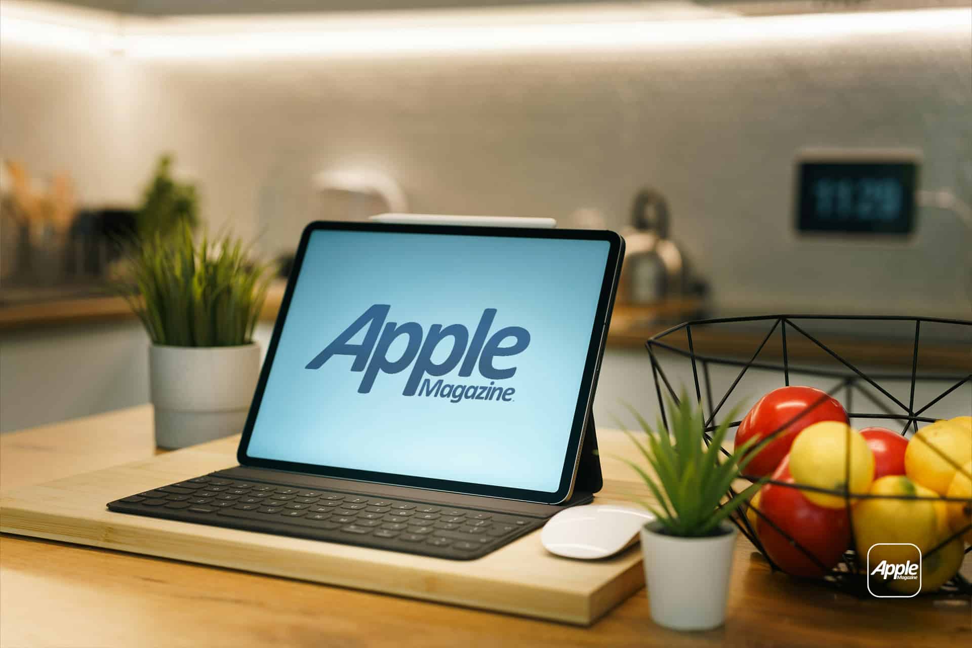 An iPad Kitchen companion with a keyboard and Apple Magazine on the screen sits on a wooden kitchen counter, surrounded by potted plants, a bowl of fruit, and kitchen appliances in the background.