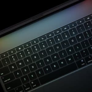 Close-up of a laptop keyboard in dim lighting, showing black keys with white letters and symbols—ideal for ipad pro coding. Function keys, a number row, and a partially visible trackpad are also captured.