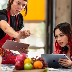 Two women use tablets at a table covered in art supplies and fruit. Their absorbed focus on the devices mirrors teenage tech preference, with one in a red apron gesturing animatedly while the other, with red hair, studies her own screen.