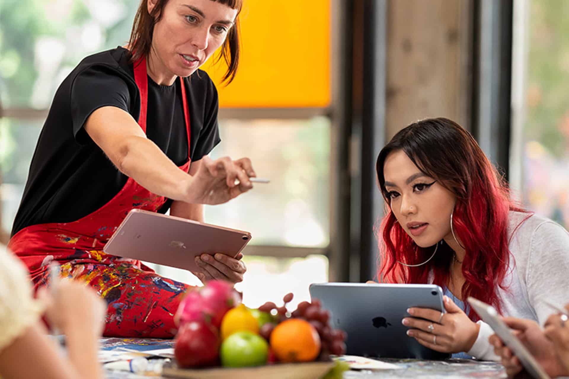 Two women use tablets at a table covered in art supplies and fruit. Their absorbed focus on the devices mirrors teenage tech preference, with one in a red apron gesturing animatedly while the other, with red hair, studies her own screen.