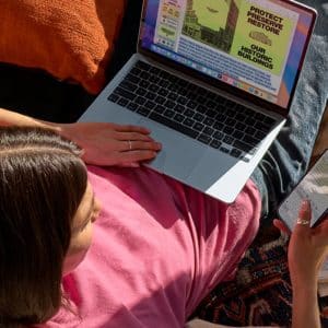 A person in a pink shirt sits on a couch using a laptop and holding a smartphone, reflecting teenage tech preference. The laptop displays a colorful page about protecting historic buildings as sunlight streams onto the cozy scene.