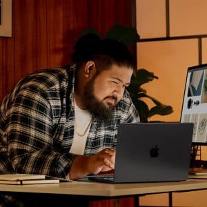 A man in a plaid shirt leans over a desk, focused on his laptop. There is a notebook nearby, and a monitor next to him displays various food images. The room has warm lighting and wooden paneling.