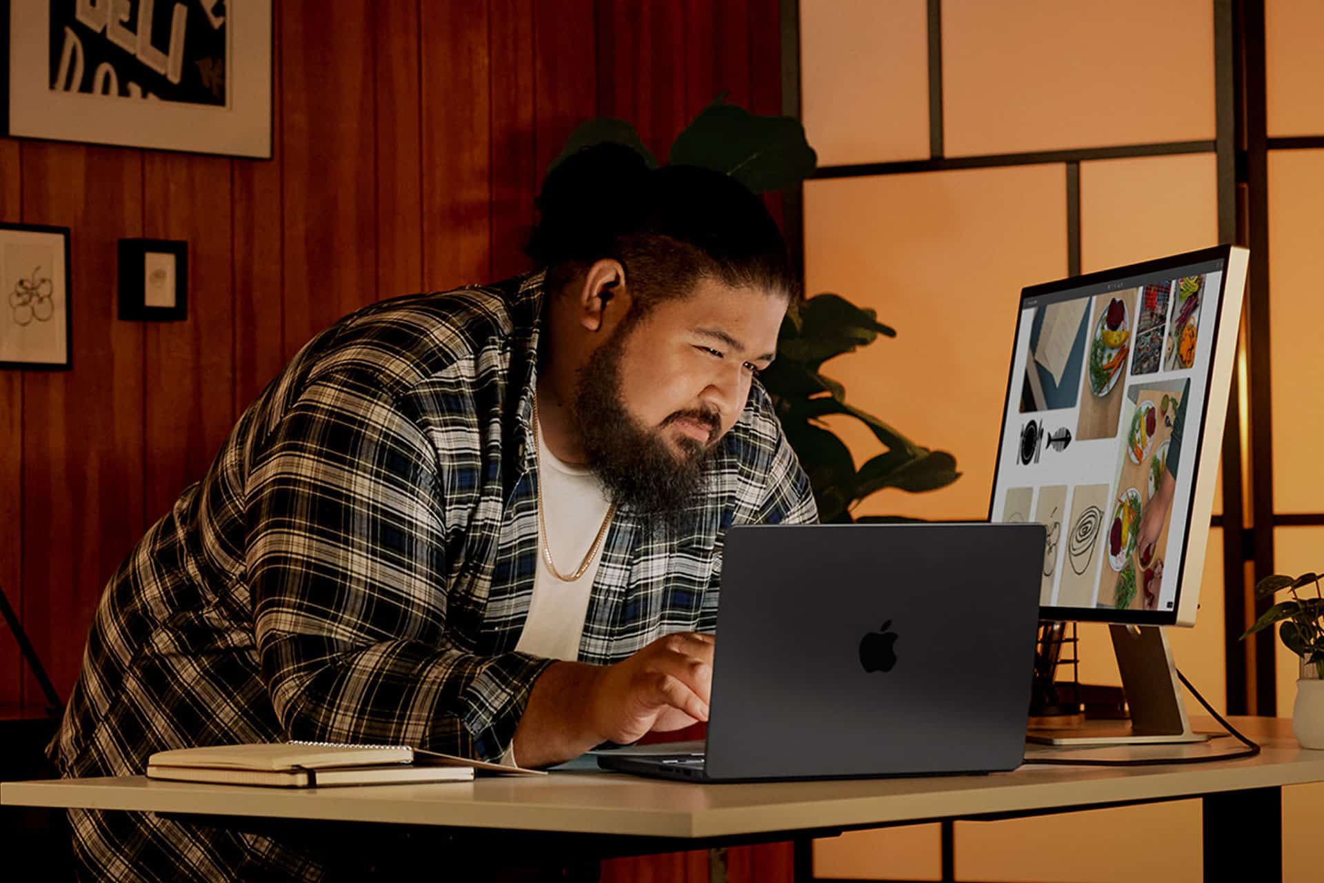 A man in a plaid shirt leans over a desk, focused on his laptop. There is a notebook nearby, and a monitor next to him displays various food images. The room has warm lighting and wooden paneling.